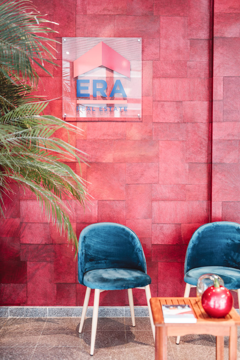 Detail van een deel van een palm in de linkerhoek, 2 blauwe velours kuipstoelen die tegen een rode muur staan met daarboven het logo van Era.